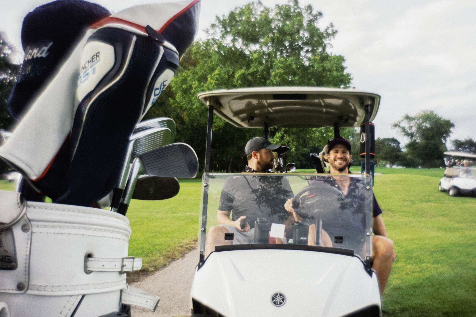 Two players grinning from a cart, framed by a bag of patterned headcovers